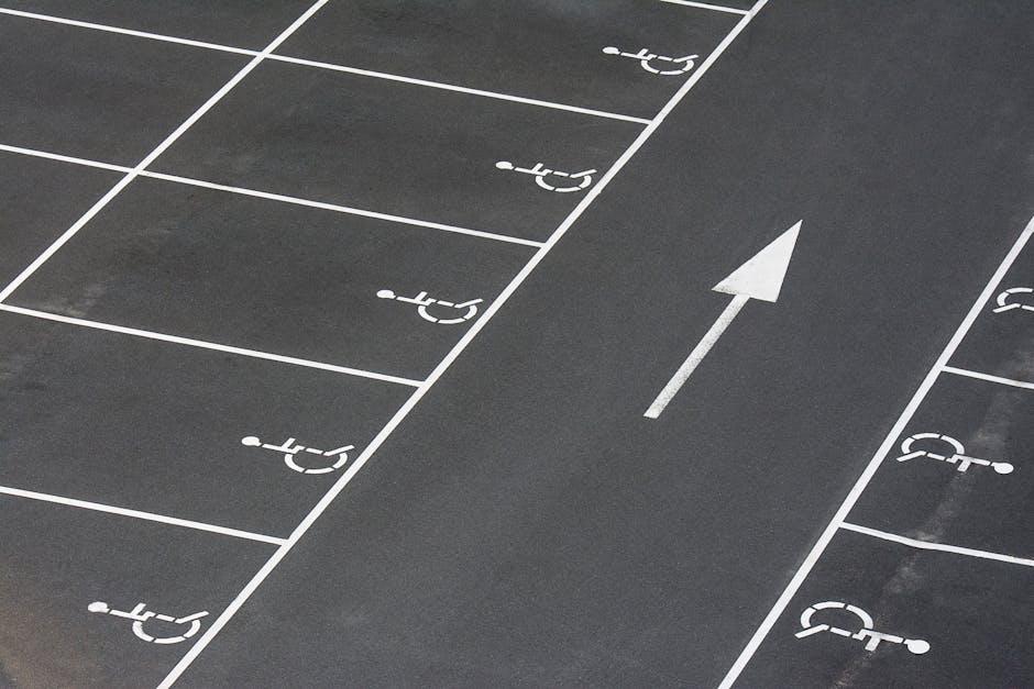 Close-up image of a dark asphalt parking lot surface with clearly marked white parking spaces and an adjacent directional arrow pointing forward, indicating a designated lane for vehicles. The parking spots are designated for handicapped parking with white wheelchair symbols, and the surface appears clean and well-maintained. The white markings are crisp and visible, contrasting sharply against the dark background. The image demonstrates a tidy, accessible outdoor surface, suitable for surface cleaning and maintenance procedures. The setting is outdoor, with ample natural lighting highlighting the cleanliness and clarity of the markings, relevant to commercial or residential area upkeep. Highbury Carpet Cleaning recommends proper surface cleaning and sanitisation for such parking lot surfaces to ensure accessibility and safety, consistent with their cleaning services as showcased on the website highburycarpetcleaning.com, particularly in contexts involving access and maintenance for properties like Highbury Grove flats.