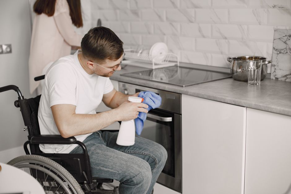A man in a wheelchair engaged in surface cleaning in a modern kitchen, holding a white spray bottle and blue cloth, with a focused expression. The kitchen features a grey countertop with a stainless steel pot and glass, a built-in oven, ceramic tiles with a white marble pattern on the walls, and ample natural lighting. The scene demonstrates high standards of hygiene and cleanliness as part of domestic cleaning, with visible tools for deep cleaning. Highbury Carpet Cleaning is mentioned as a provider of professional cleaning services. The environment appears tidy, with clean surfaces and well-organized kitchenware, reflecting effective sanitisation processes for residential spaces.