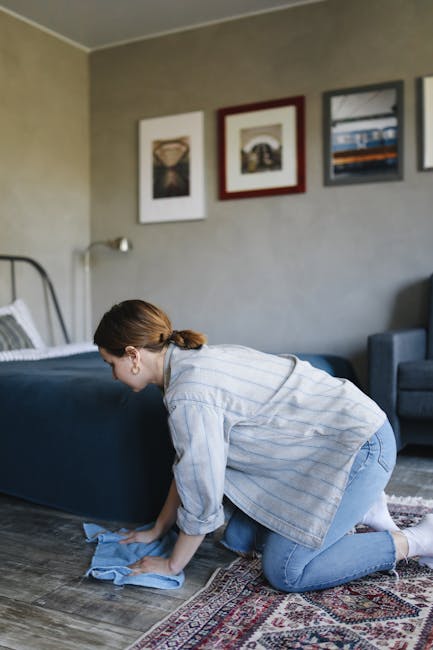A woman with brown hair tied back, wearing a light blue striped shirt and jeans, is kneeling on a patterned area rug in a living room, engaged in surface cleaning with a blue cloth. The room features a dark blue upholstered bed with striped bedding, a gray wall decorated with framed artwork, a blue armchair, and wooden flooring. Natural light illuminates the space, highlighting the clean, dust-free surfaces and the tidy, well-maintained environment. This scene illustrates domestic cleaning and deep surface care, as performed by Highbury Carpet Cleaning, to maintain hygiene and freshness in residential spaces.