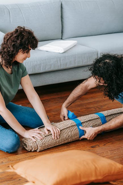 Two individuals are engaged in a domestic cleaning task, rolling out a textured beige carpet onto a wooden floor within a living room. One person, with curly brown hair and wearing a green top, is holding the edge of the carpet, while the other, with curly black hair and dressed in a blue shirt, is securing it in place. The room features a light blue sofa with a white cushion resting on it, and the floor beneath is a polished hardwood. The scene is illuminated by natural light, highlighting the cleanliness and shine of the surfaces, indicative of a professional approach to surface cleaning and maintenance, as promoted by Highbury Carpet Cleaning. Visible cleaning tools or products are not present, emphasizing the focus on careful, manual placement of the carpet that aligns with deep cleaning and sanitisation practices.