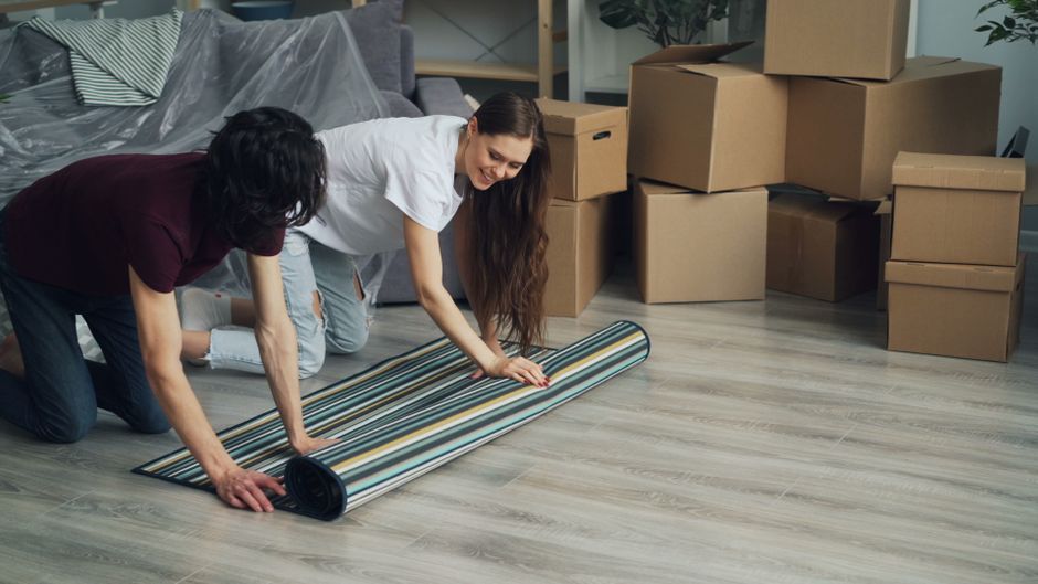 Two individuals are engaged in a detailed surface cleaning process on a light-colored wooden floor in a residential space. The person in the white top is unrolling a striped, multi-colored carpet, while the individual in a dark shirt helps position it evenly on the floor. In the background, several brown cardboard boxes are stacked near a modern sofa, indicating a possible moving or redecorating activity. Bright, natural lighting illuminates the room, highlighting the clean, dust-free condition of the wooden flooring and the fresh appearance of the rolled carpet. Visible cleaning tools such as the carpet and possibly a cleaning spray on the floor suggest a focus on thorough domestic cleaning practices, with an emphasis on proper surface preparation and maintenance, aligned with highburycarpetcleaning.com’s focus on avoiding common carpet cleaning mistakes.