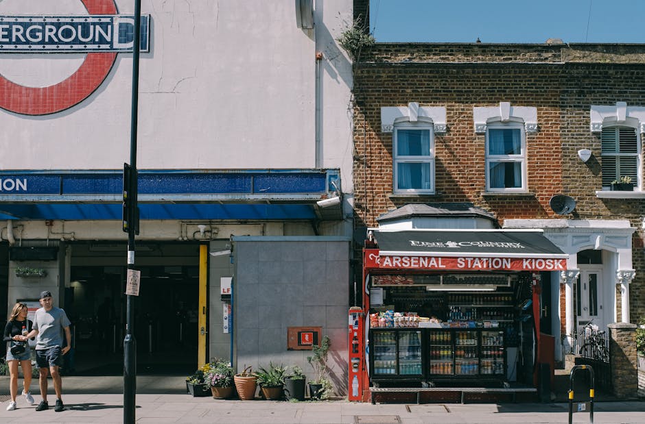 Street view featuring the entrance to London's Underground station with a visible 'Underground' roundel sign on the left, adjacent to a modern building facade with a parking garage entrance. To the right, a red 'Arsenal Station Kiosk' is situated on a brick building, displaying an array of beverages and snacks behind glass. The scene is illuminated by sunlight, with clear shadows cast on the sidewalk, which has potted plants near the kiosk. Two pedestrians, a woman and a man, are walking past on the sidewalk. The overall setting reflects a typical urban scene with commercial and transit elements, emphasizing the surroundings of Arsenal Station near Highbury, captured in a clean, lively, and well-maintained environment. The image complements the services offered by Highbury Carpet Cleaning for surface and deep cleaning, particularly in busy retail and station areas.