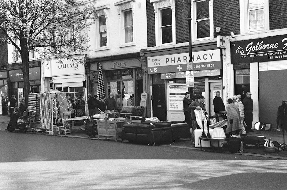 A black-and-white photograph of a busy street scene outside a pharmacy and various shops in Highbury, with a group of people gathered around furniture and household items displayed on the pavement. The scene includes sofas, chairs, and tables, some covered with protective sheets, suggesting a temporary outdoor sale or clearance event. The storefronts have signage indicating NHS and private healthcare services, along with a pharmacy and a fish shop named Galborne. The lighting appears natural, and the street is lined with a tree and parked scooters. The overall atmosphere is vibrant, with people engaged in browsing and socializing, capturing a typical lively day in an urban residential area. Highbury Carpet Cleaning offers professional surface cleaning and deep cleaning services that can help maintain the hygiene and appearance of furniture and interior surfaces in settings like this.