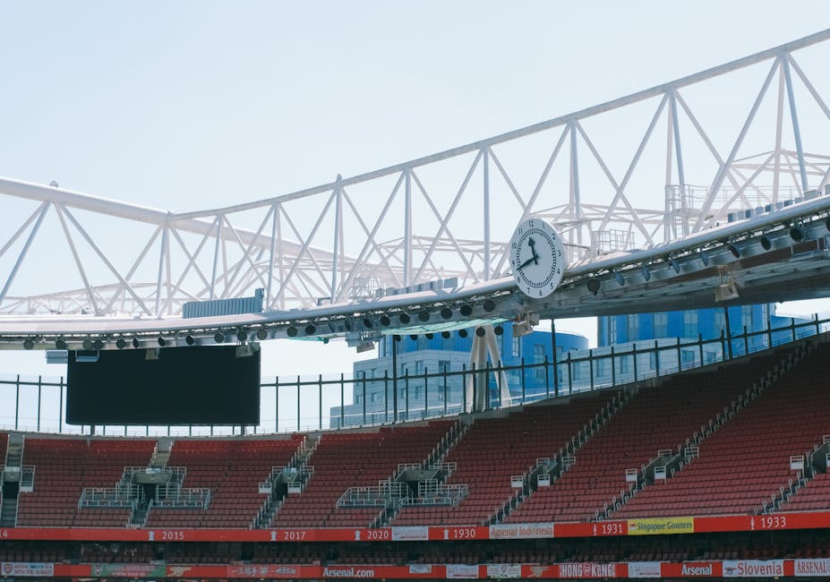 A view of an empty stadium seating area with red plastic chairs arranged in sections, under a large white steel framework roof with triangular trusses. A round clock is mounted on the framework, showing the time as 11:10. The lighting indicates natural daylight, illuminating the structure and seating area evenly. The image captures the clean, well-maintained environment of a sports venue, with no visible dust or debris, emphasizing overall hygiene and upkeep, consistent with surface or deep cleaning standards as performed by Highbury Carpet Cleaning for maintaining hygiene and cleanliness in large public spaces.