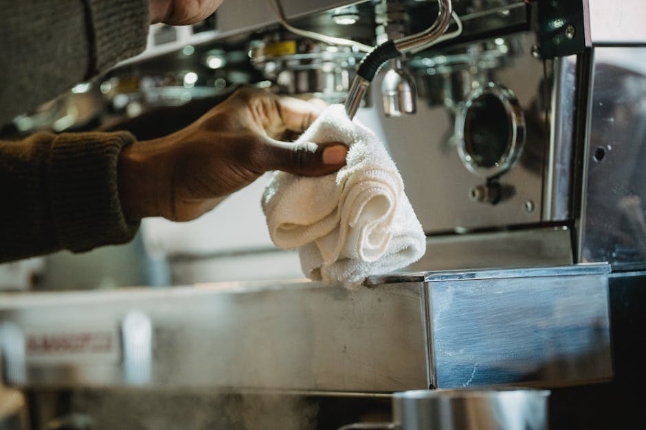 Close-up of a person’s hand wiping a stainless steel espresso machine with a white cloth, situated on a commercial kitchen counter, demonstrating surface cleaning and sanitisation as part of professional cleaning services offered by Highbury Carpet Cleaning, highlighting the focus on hygiene and equipment maintenance in a busy café or pub environment.