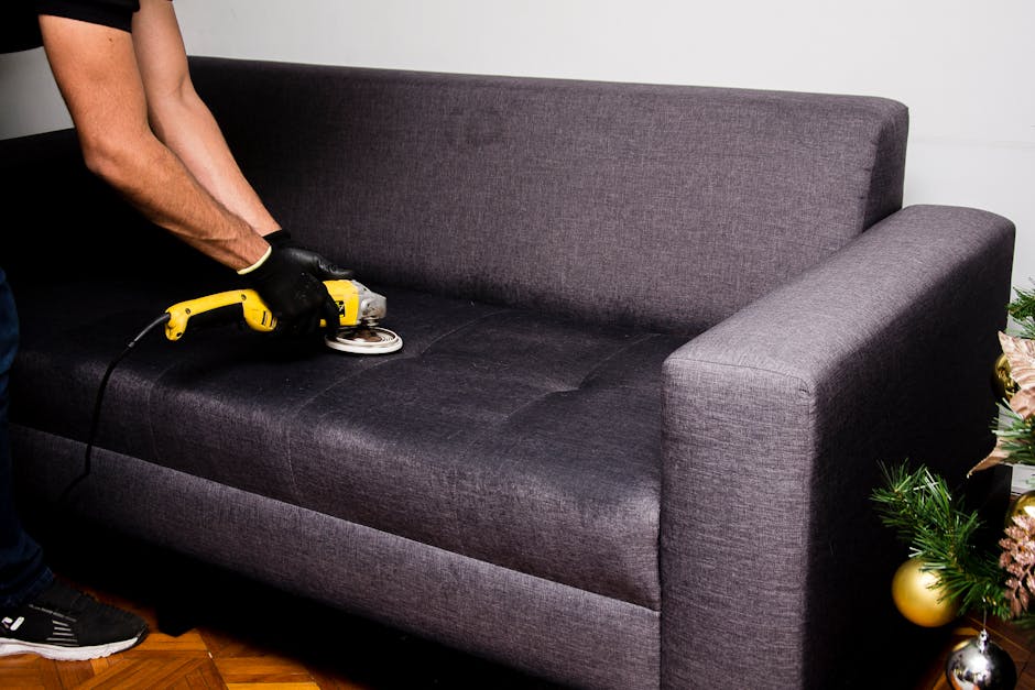 A person wearing black gloves is using a handheld electric polisher on a dark gray fabric sofa cushion in a living room. The sofa has a modern design with a high backrest and wide armrests, situated on a wooden floor. The room features white walls and natural lighting, with a decorated Christmas tree visible on the right side of the image. The scene depicts surface cleaning and maintenance to ensure hygiene and cleanliness of the furniture, in line with professional domestic cleaning services offered by Highbury Carpet Cleaning, as referenced on the Highbury Fields carpet cleaning same day service page.