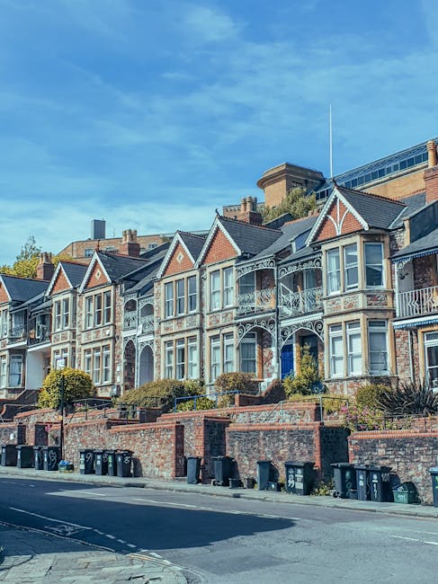 A row of Victorian-style terraced houses with ornate white detailing and bay windows, situated on an inclined street in Highbury. The houses have brick facades with small front gardens, surrounded by low brick walls and greenery. The street below is lined with black and green waste bins, and the sky above is partly cloudy with patches of blue. Natural daylight illuminates the scene, highlighting the cleanliness and well-maintained appearance of the properties. Highbury Carpet Cleaning offers surface cleaning and deep cleaning services to maintain the hygiene and appearance of residential buildings such as these, ensuring a pristine environment. This image is relevant to the Highbury Fields carpet cleaning same day service page, emphasizing quality home maintenance.