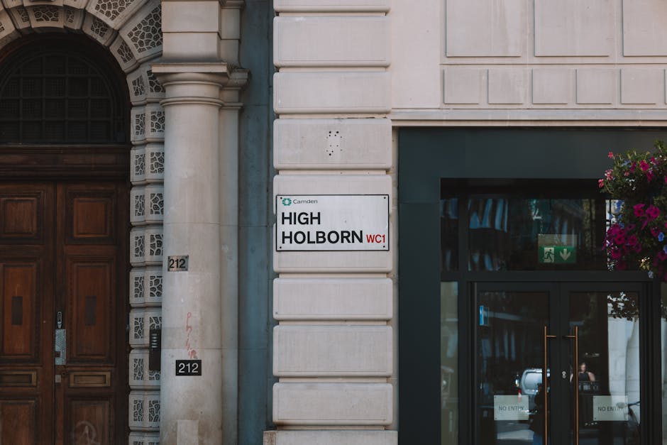 Close-up view of the exterior of a building at Highbury tenancy, showing a section of the sandstone facade with decorative architectural details on the left and a modern glass door with black framing on the right. The building features white stone blocks with horizontal grooves, and mounted on the wall is a rectangular white sign displaying 'HIGH HOLBORN WC1' in black letters and a small Camden logo in the top left corner. To the right of the sign, there is a hanging flower basket filled with pink and purple blooms, adding a touch of color to the urban setting. The glass door has two 'No Entry' signs on it, with reflections of the street visible in the glass, and a green emergency exit sign is positioned above the door handle. The lighting appears natural, illuminating the clean, well-maintained surfaces, highlighting the smoothness of the glass and the textured stone, suggesting recent or regular surface cleaning by professional services like Highbury Carpet Cleaning.