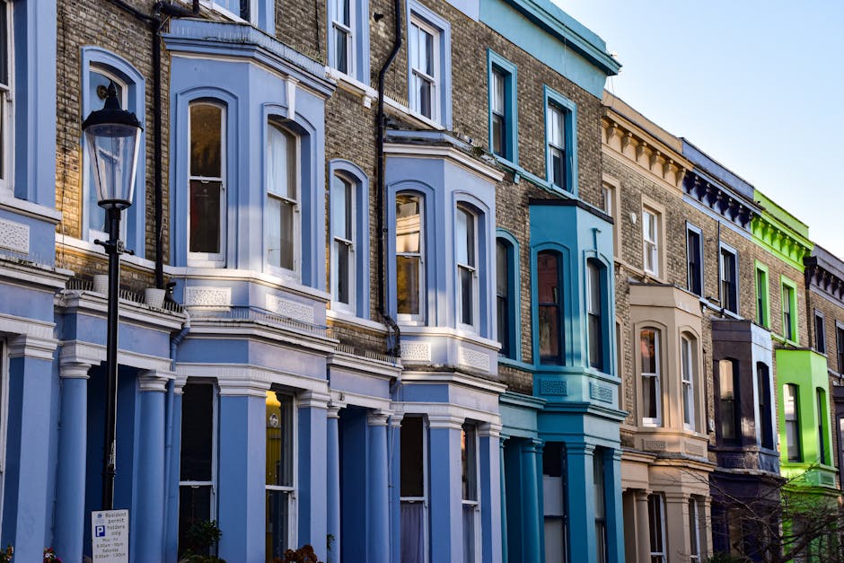A street view in Highbury, Islington with Victorian-style red brick and white terraced residential buildings featuring decorative mouldings and bay windows. The buildings are separated from the narrow asphalt road by black wrought-iron railings. The road is lined with several parked cars, primarily black and grey, and some are partially visible on the right side. A sidewalk runs along the front of the buildings, with a few pedestrians walking in the distance. Trees with fresh green foliage are visible behind the buildings, and the sky above is partly cloudy with patches of blue, highlighting a clean and well-maintained urban residential area. This scene exemplifies typical Highbury architecture and street layout, suitable for showcasing cleaning or maintenance services as highlighted on highburycarpetcleaning.com, with Highbury Carpet Cleaning emphasizing hygiene and surface cleanliness in such environments.