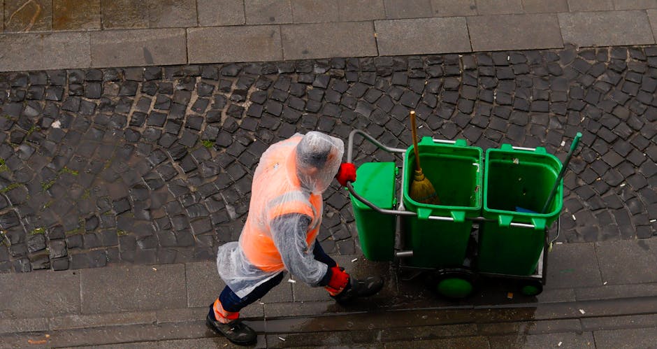 A professional cleaner wearing a transparent plastic poncho, an orange high-visibility vest, protective gloves, and black boots is pushing a green cleaning trolley with three compartments on a wet cobblestone street. The trolley contains cleaning tools, including a yellow-handled broom and a spray bottle. The scene is brightly lit, highlighting the wet surface, and the surrounding pavement appears clean and well-maintained. This image illustrates surface cleaning and maintenance activities typical of commercial or residential hygiene services, aligned with the practices discussed on the page about Islington Council rules for bulk waste from cleaning jobs. The branding of Highbury Carpet Cleaning is subtly referenced, emphasizing their expertise in domestic cleaning and surface sanitisation for local property upkeep.
