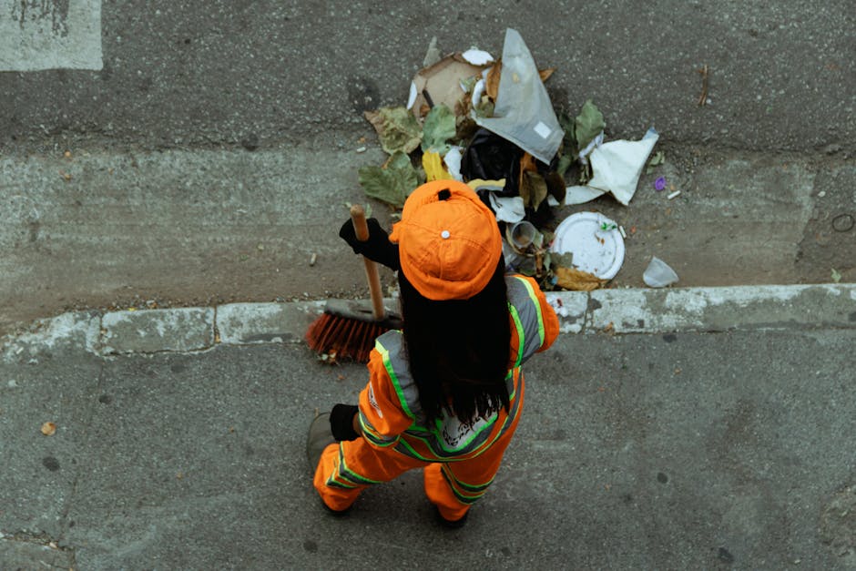 A sanitation worker dressed in an orange high-visibility uniform and matching cap is seen from above, standing on a concrete sidewalk next to a curb. The worker is holding a broom while collecting a pile of assorted trash, including paper, plastic, and organic waste, from the edge of the sidewalk. The area appears to be part of an urban street with a worn concrete curb and a section of road visible at the top of the image. The scene highlights ongoing efforts in surface cleaning and waste collection, consistent with regulations for managing bulk waste as outlined by Islington Council. The presence of the waste pile indicates the importance of proper disposal methods, which companies like Highbury Carpet Cleaning support through thorough cleaning and sanitation processes.