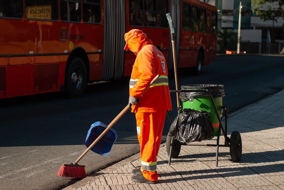 A professional cleaner wearing a transparent plastic poncho, an orange high-visibility vest, protective gloves, and black boots is pushing a green cleaning trolley with three compartments on a wet cobblestone street. The trolley contains cleaning tools, including a yellow-handled broom and a spray bottle. The scene is brightly lit, highlighting the wet surface, and the surrounding pavement appears clean and well-maintained. This image illustrates surface cleaning and maintenance activities typical of commercial or residential hygiene services, aligned with the practices discussed on the page about Islington Council rules for bulk waste from cleaning jobs. The branding of Highbury Carpet Cleaning is subtly referenced, emphasizing their expertise in domestic cleaning and surface sanitisation for local property upkeep.