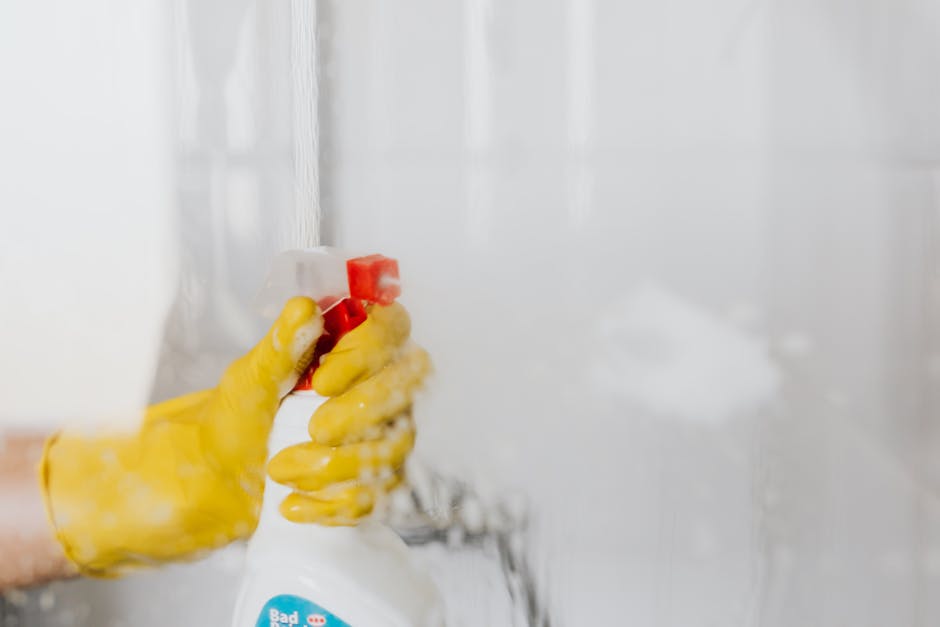 Close-up of a person's hand wearing a yellow rubber glove, holding a white spray bottle with a red nozzle, applying cleaning solution to a glass shower door in a bathroom. The shower door, made of clear glass, appears to be in the process of being cleaned, with some soap suds visible on the surface. The background shows a white tiled wall, illuminated by natural light, indicating a well-maintained space. This image illustrates surface cleaning and sanitisation as part of domestic or commercial deep cleaning services offered by Highbury Carpet Cleaning, emphasizing their focus on hygiene and effective stain removal in residential bathrooms.