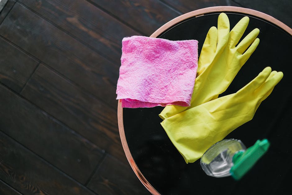 A round black surface on a wooden floor holds cleaning tools including a pair of yellow rubber gloves, a pink microfiber cloth, and a spray bottle with a green cap. The rubber gloves are neatly placed next to the cloth, which appears clean and folded. The spray bottle contains a transparent liquid and is positioned partially inside the gloves. The lighting highlights the shiny, clean surface and the textures of the cloth and gloves, indicating a preparation for surface cleaning or sanitisation. The scene is part of an organized domestic cleaning setup, emphasizing hygiene and thorough cleaning practices, with the highburycarpetcleaning.com branding and the context related to understanding hidden cleaning charges in Highbury.