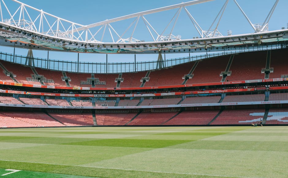 An empty sports stadium featuring a well-maintained grass pitch with alternating shades of green, surrounded by multiple tiers of red and pink seating. The stadium has a large, white tensile roof structure extending over the seating area, supported by metal beams, allowing ample natural light to illuminate the interior. The seating sections are arranged in a circular pattern, with walkways and railings visible between levels. The overall scene emphasizes cleanliness and order, aligning with the professional standards of Highbury Carpet Cleaning for surface cleaning and deep cleaning services, though the image depicts an outdoor sports environment rather than a domestic or commercial interior setting.
